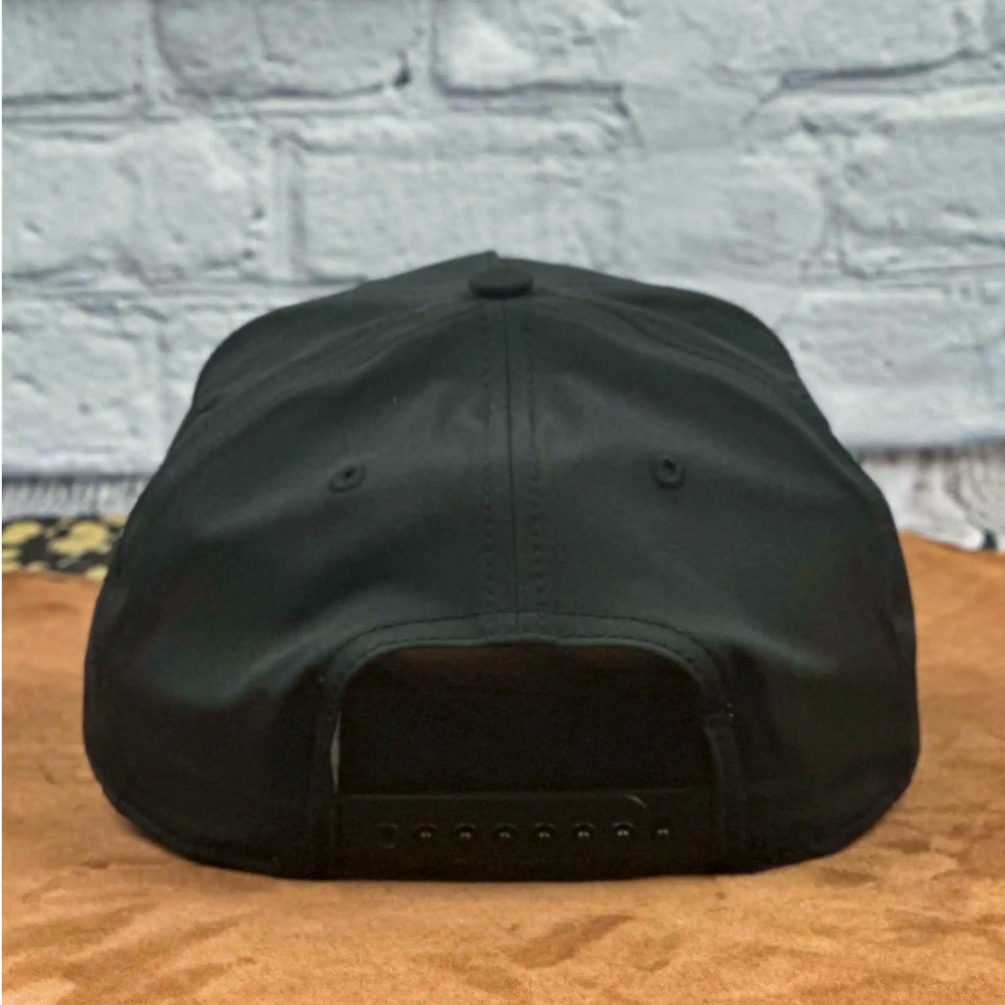 Rear view of a black Richardson 258 five-panel snapback hat showcasing the adjustable snap closure, set on a tan surface with a white brick wall background.