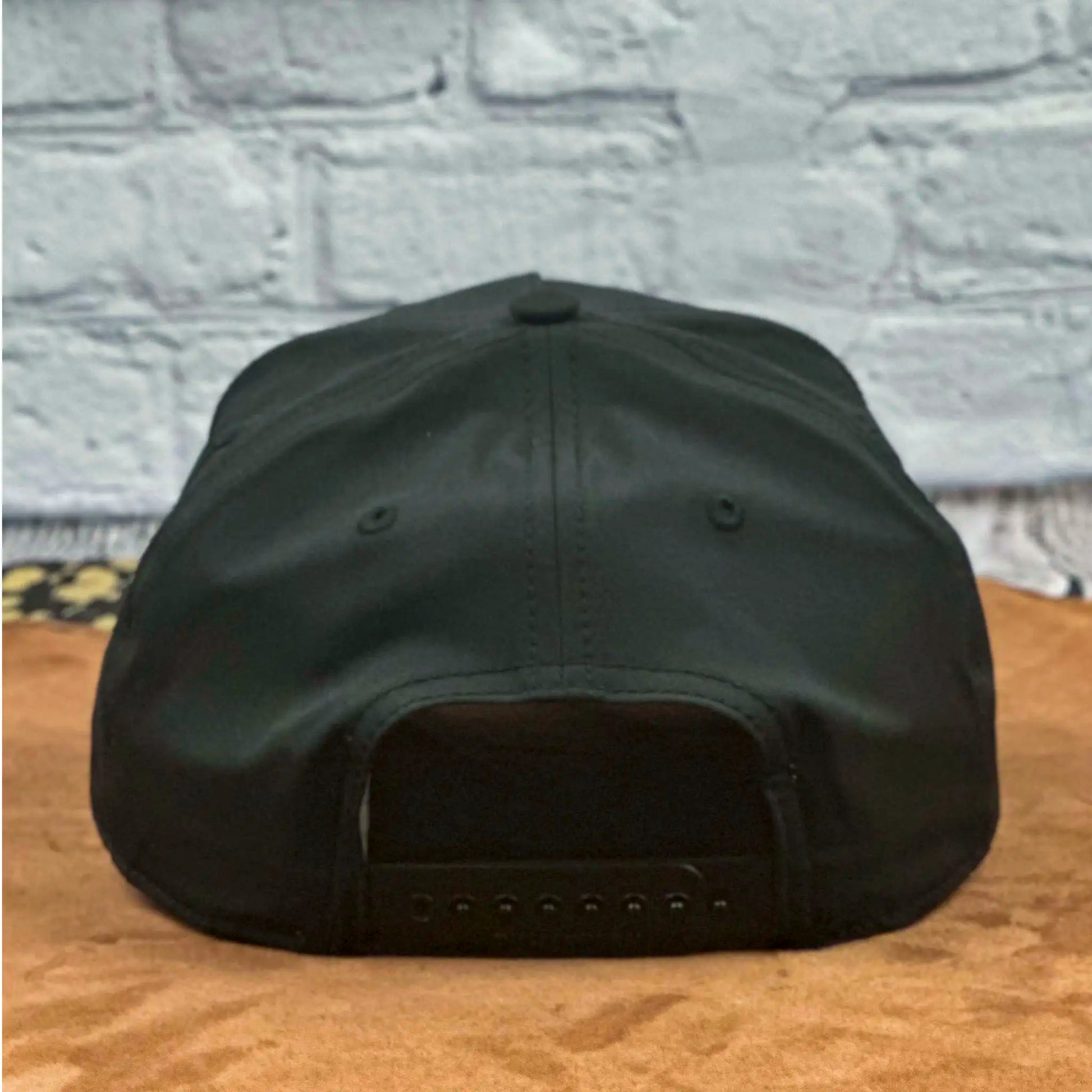 Rear view of a black Richardson 258 five-panel snapback hat showcasing the adjustable snap closure, set on a tan surface with a white brick wall background.