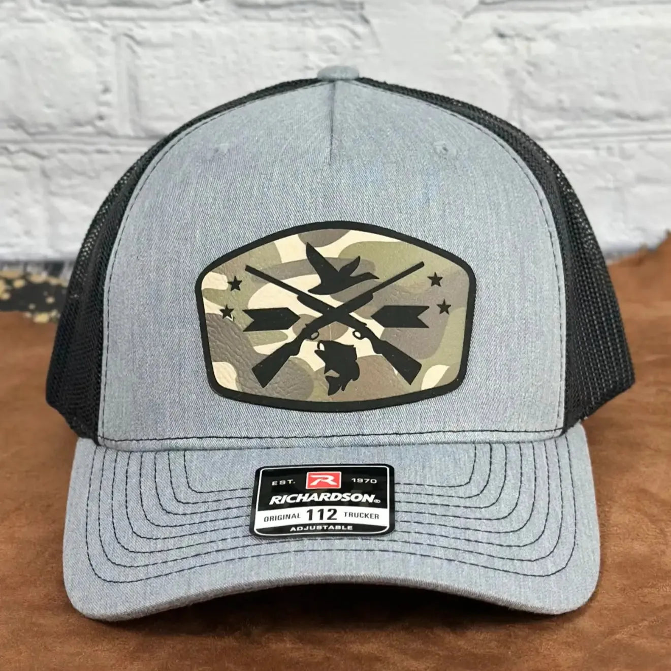 Front view of a grey and black Richardson 112 adjustable trucker hat featuring a camo patch with crossed rifles, duck, dog, and stars design, displayed on a tan surface with a white brick background.