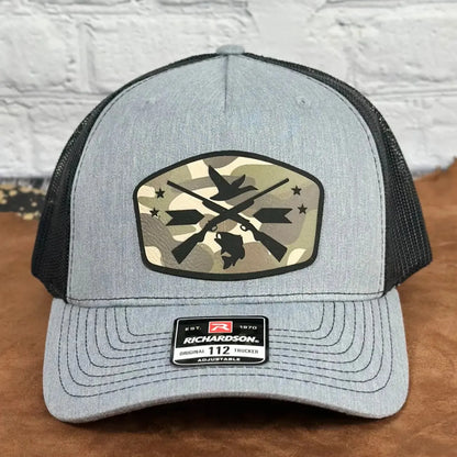 Front view of a grey and black Richardson 112 adjustable trucker hat featuring a camo patch with crossed rifles, duck, dog, and stars design, displayed on a tan surface with a white brick background.