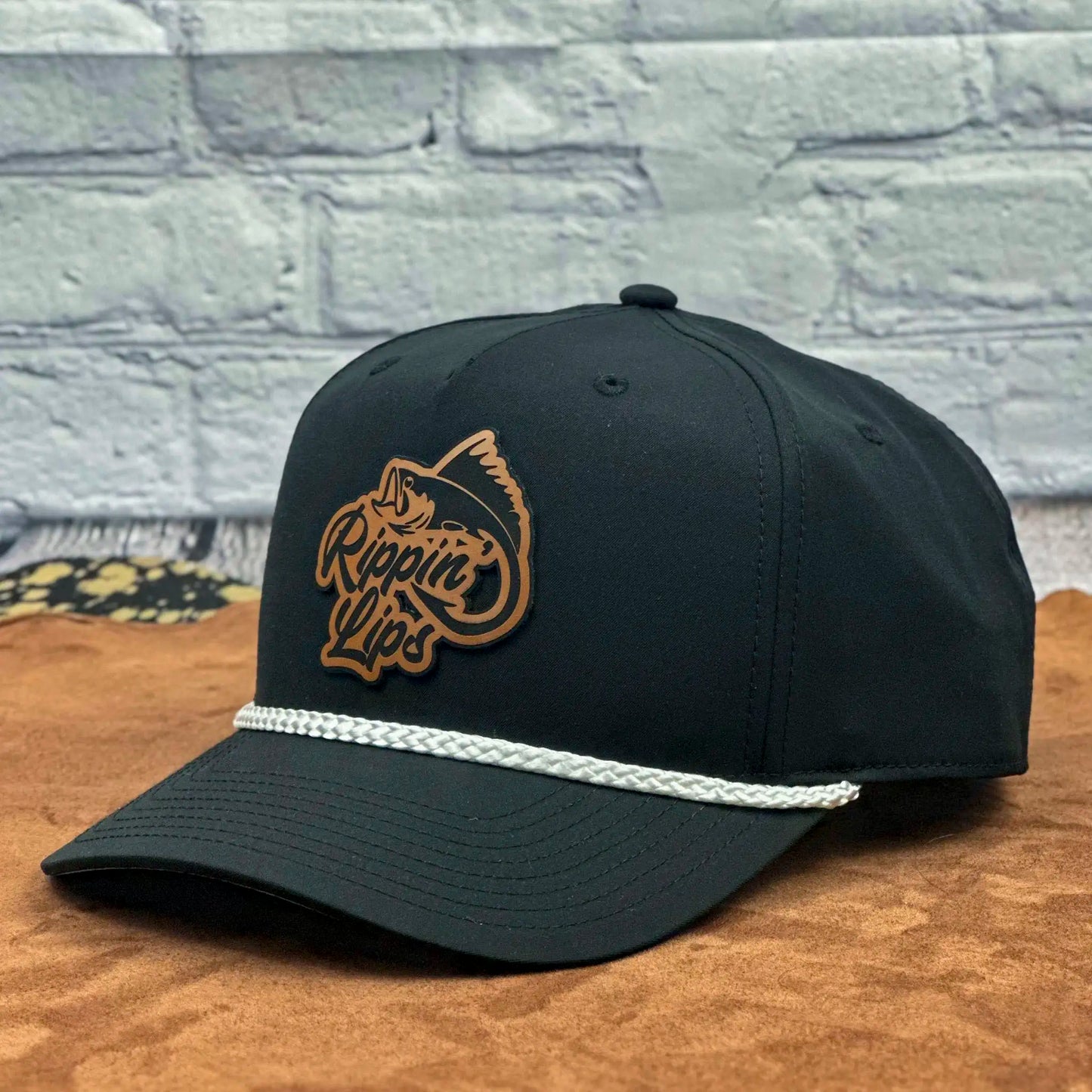Angled view of a black Richardson 258 five-panel rope hat featuring a brown 'Rippin' Lips' leather fish logo patch and white braided rope across the brim, displayed on a tan surface with a light brick wall background.