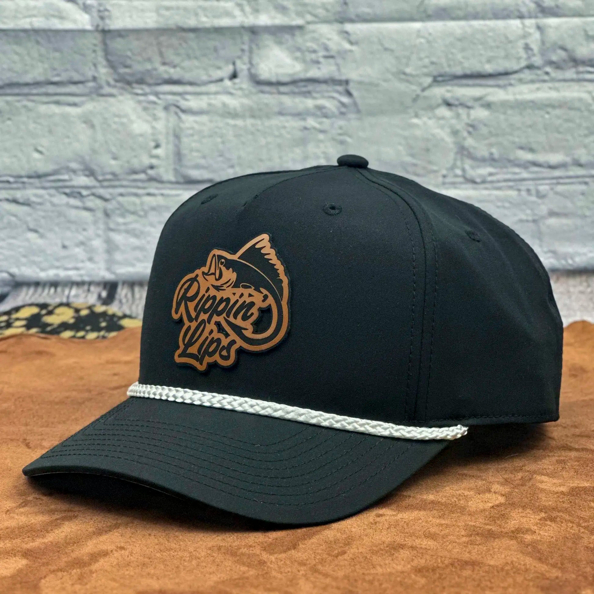 Angled view of a black Richardson 258 five-panel rope hat featuring a brown 'Rippin' Lips' leather fish logo patch and white braided rope across the brim, displayed on a tan surface with a light brick wall background.