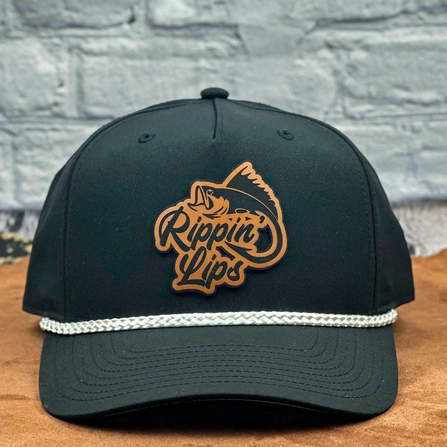 Black Richardson 258 five-panel snapback hat featuring a brown leather 'Rippin' Lips' fish logo patch and a white braided rope detail across the brim, displayed on a brown surface with a brick background.
