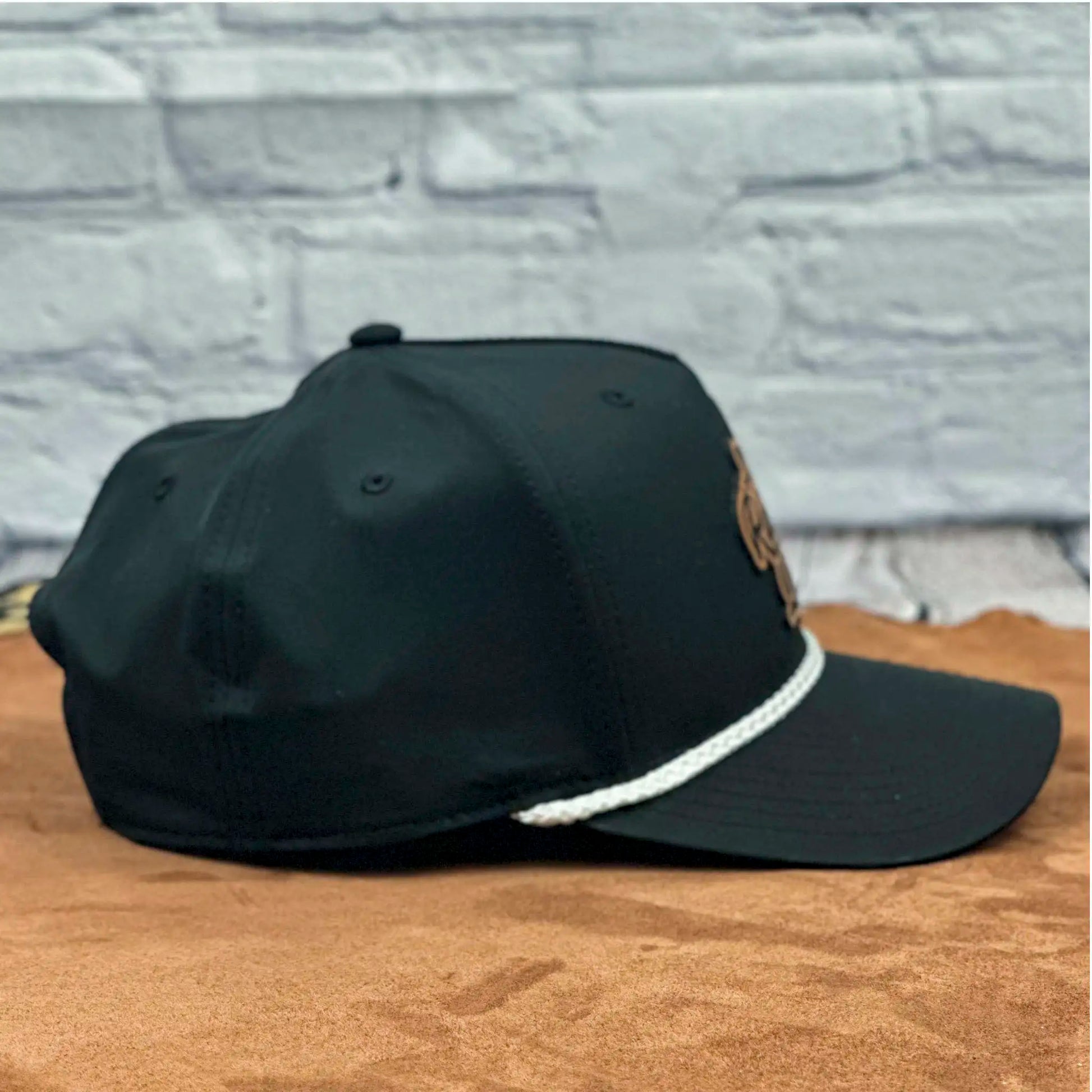 Side profile of a black Richardson 258 five-panel snapback hat with a white braided rope detail along the brim, showing the clean and structured panel design on a tan surface with a light brick wall background.