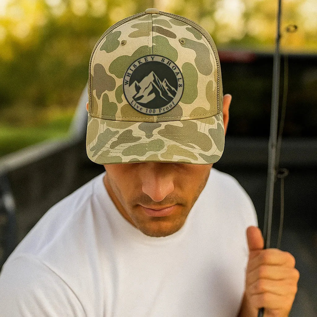 Man wearing a camo Richardson-style six-panel snapback hat with a black 'Whiskey Shoals – Live 100 Proof' mountain logo patch, holding a fishing rod next to a truck in an outdoor setting.